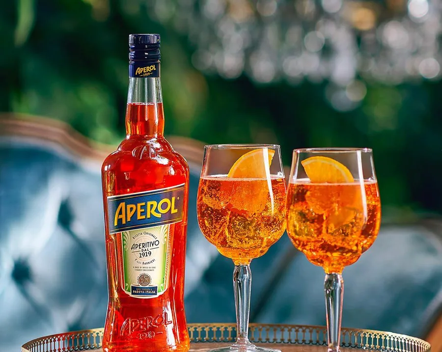 Aperol Spritz em taça com rodela de laranja, ao lado da garrafa de Aperol